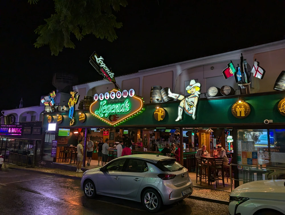 The lively nightlife on the main bar strip in Albufeira, featuring the brightly lit, western-themed "Legends Bar" at night.