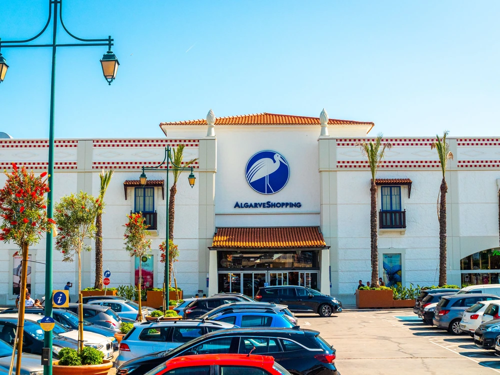 The modern entrance to Algarve Shopping, a popular mall in Albufeira, with palm trees and a busy car park on a sunny day.
