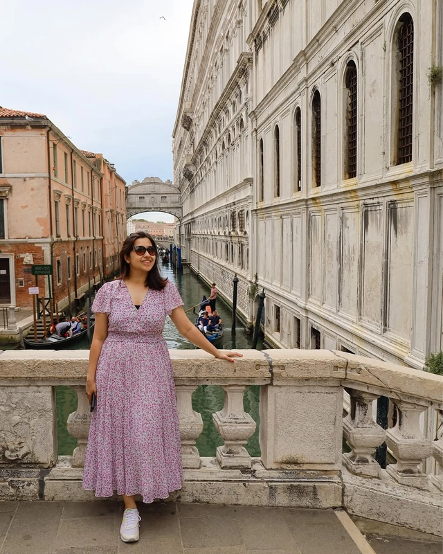 A woman in a floral dress stands on a bridge overlooking a canal, with the famous enclosed Bridge of Sighs visible in the background in Venice.