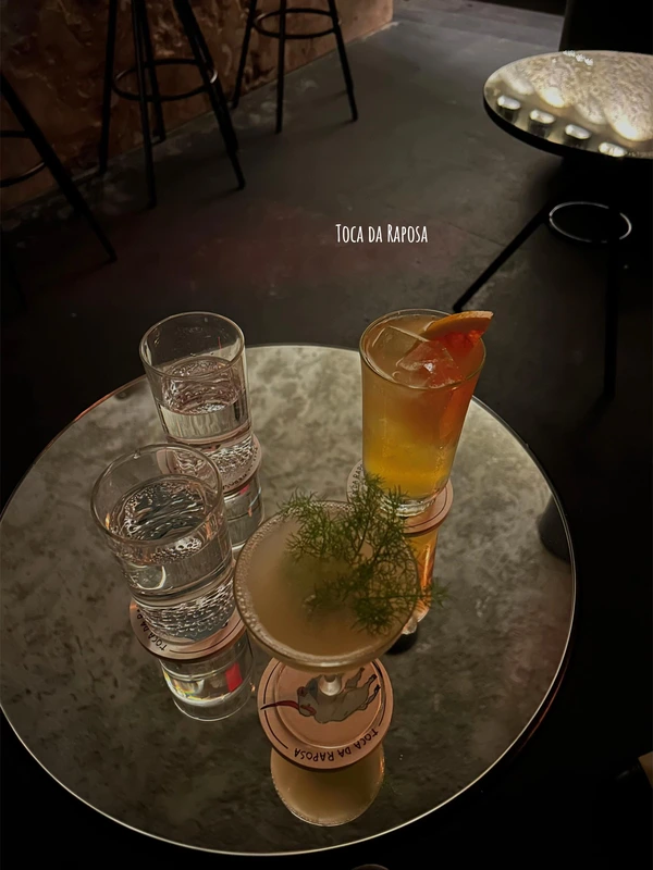 Creative cocktails, including one with a fresh dill garnish, served on a reflective table at Toca da Raposa cocktail bar in Lisbon.