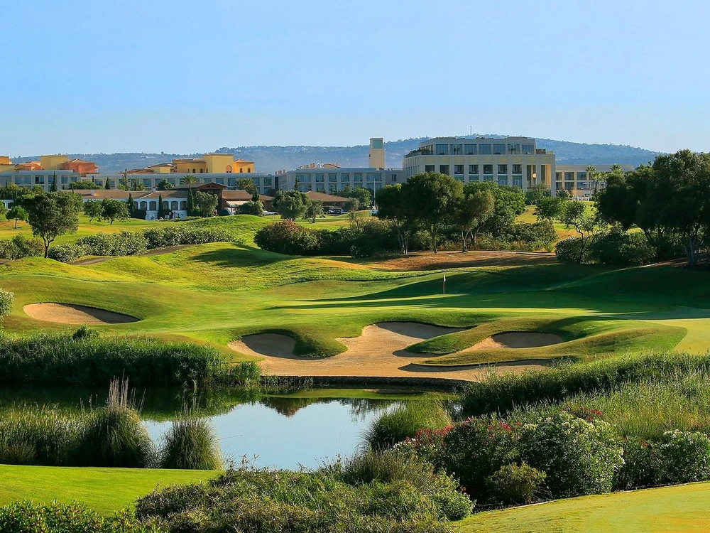 A scenic view of the Dom Pedro Victoria Golf Course in Vilamoura, featuring rolling hills, a water hazard, and resort hotels in the background.
