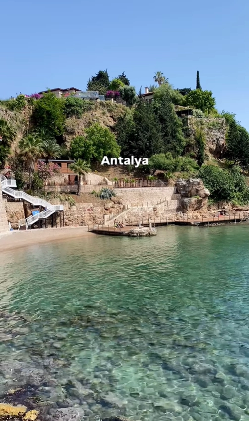 A beautiful secluded beach with clear turquoise water nestled at the base of a lush green cliff in Antalya, Turkey.