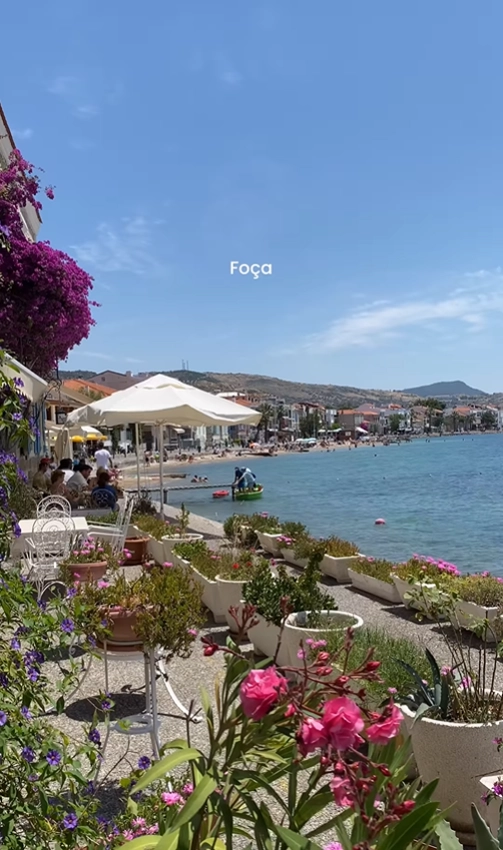A picturesque view of the harbor in Foça, Turkey, featuring vibrant flowers and waterfront cafes along the Aegean coast.