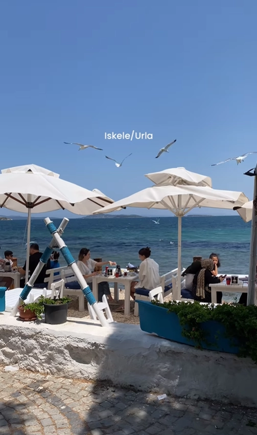 Diners relaxing at a lively beach cafe in İskele, Urla, Turkey, enjoying the seaside view under large white umbrellas.
