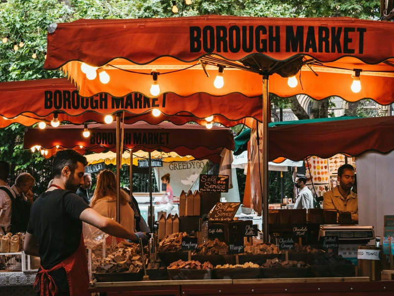 A vendor at a food stall with a red awning at Borough Market, one of the best markets in London, serving artisanal sweets to customers.