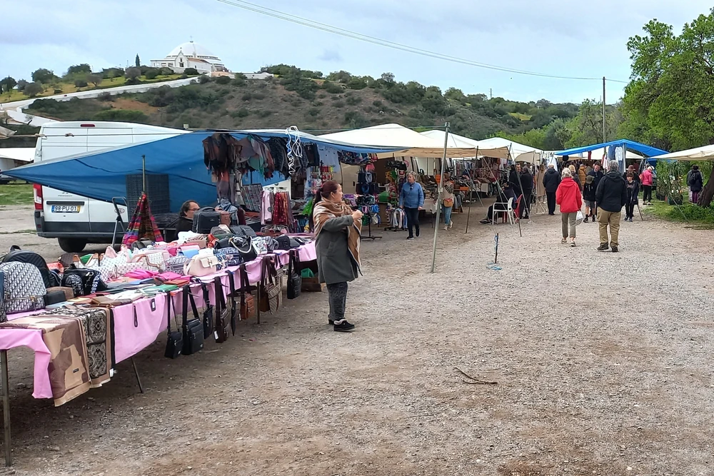 The bustling outdoor Loulé Gypsy Market (Feira da Loulé), with vendors selling various goods under tents and a white church visible on the distant hill.