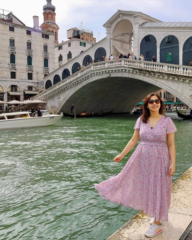A woman in a floral dress stands on the edge of the Grand Canal with the historic Ponte di Rialto bridge in the background in Venice, Italy.