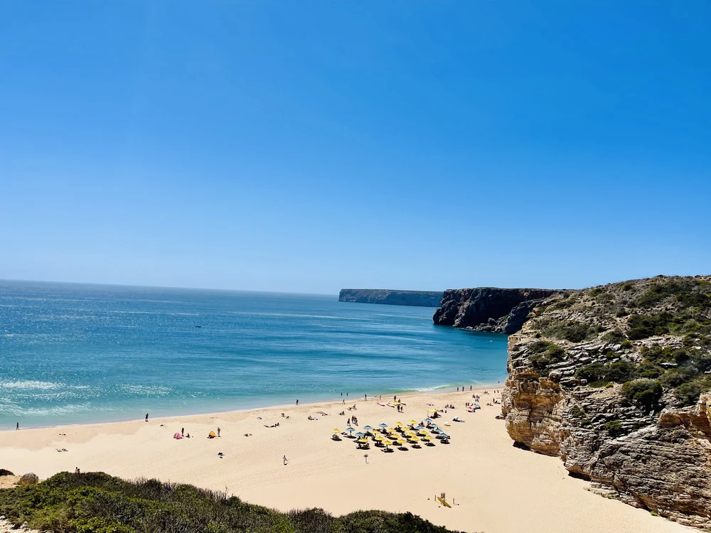 Praia do Beliche in Sagres with blue waters and yellow sun umbrellas dotting the shoreline beneath the cliffs.