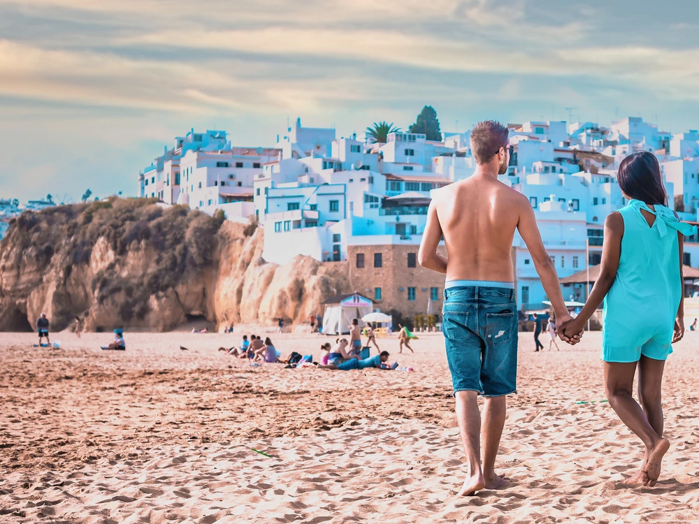 A romantic scene of a couple holding hands while walking on the golden sands of Praia dos Pescadores, with the stunning Albufeira cityscape in the background.