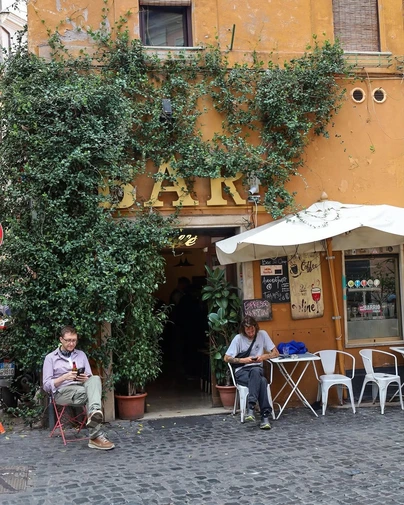 A charming, ivy-covered "BAR" on a quiet, cobblestoned street embodies the experience of discovering hidden gems and non-touristy things in the local neighborhoods of Rome.