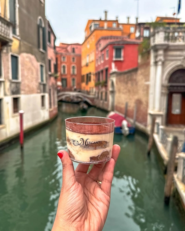 A hand holds a cup of tiramisu from I Tre Mercanti in front of a picturesque canal with colorful buildings in Venice.