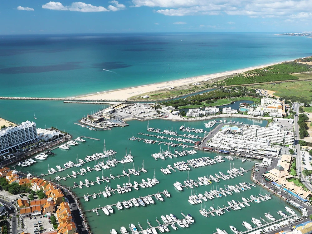 Sweeping aerial view of the Vilamoura Marina during the day, showing the turquoise harbor, luxury yachts, and the long sandy beach.