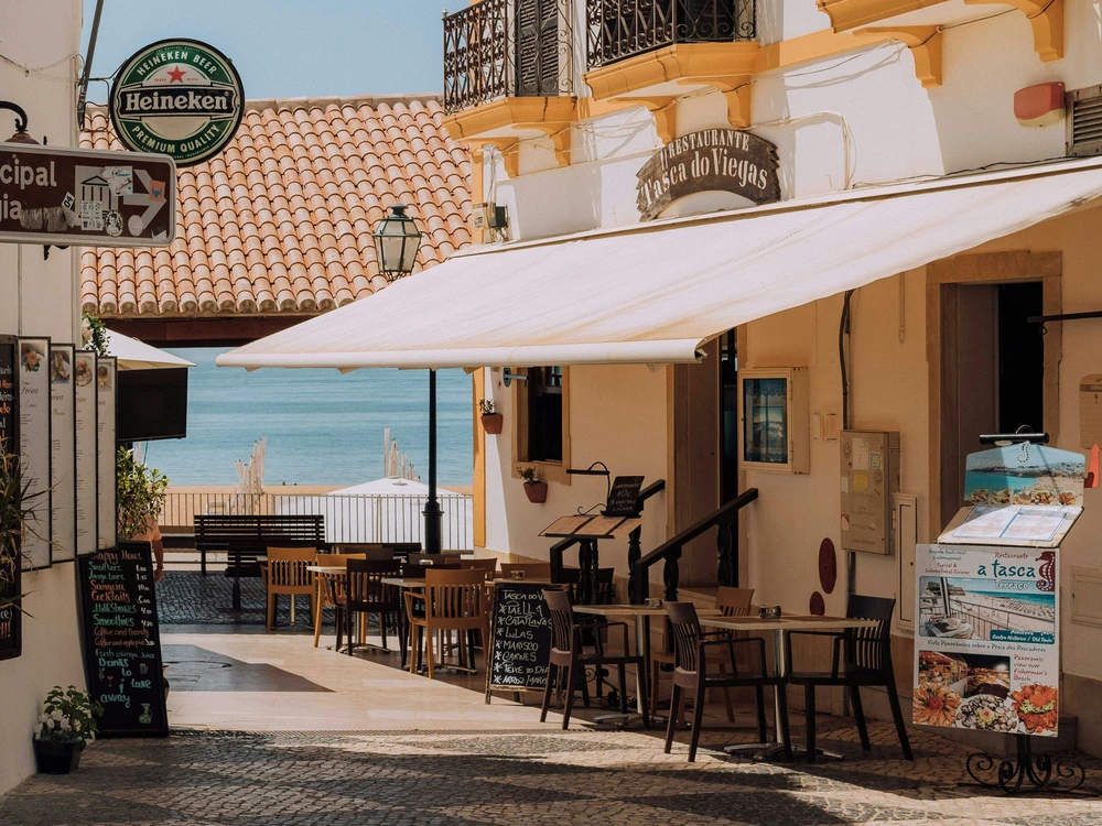 The charming Tasca do Viegas restaurant in Albufeira, Portugal, with its inviting outdoor seating on a narrow street offering a beautiful view of the ocean.