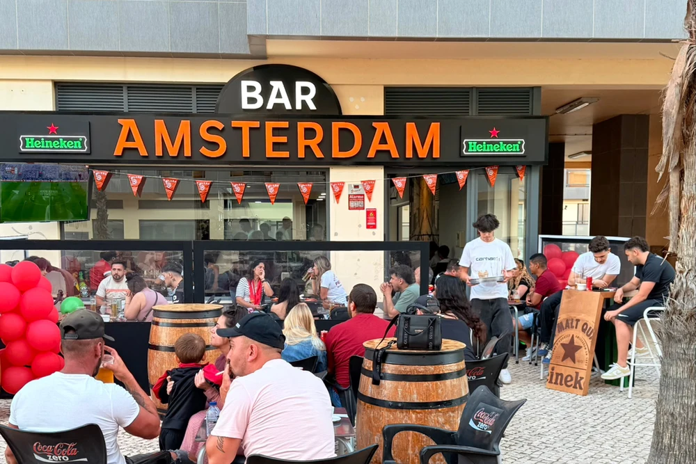 Exterior seating area of Bar Amsterdam with people sitting at barrel tables and the prominent sign overhead in Olhao, Portugal.