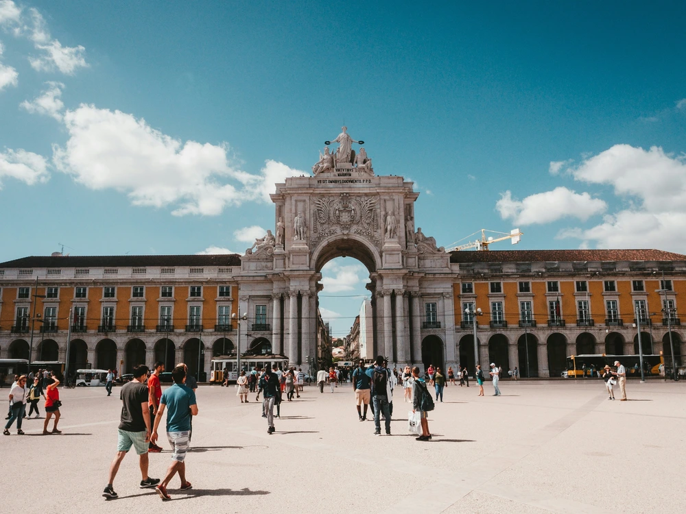 A bustling sunny day at the expansive Praça do Comércio square in Baixa, Lisbon, with tourists walking towards the majestic Rua Augusta Arch.
