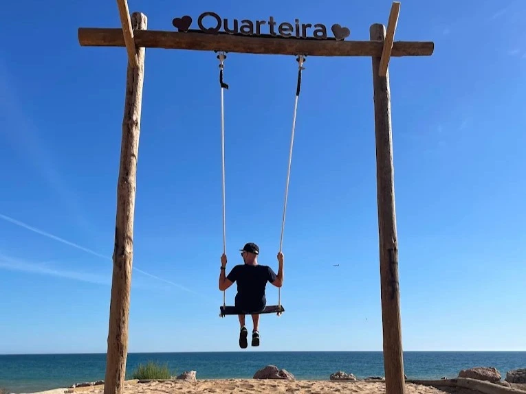 A man on the iconic Baloiço da Praia da Gaivota wooden swing overlooking the ocean in Quarteira, Portugal.
