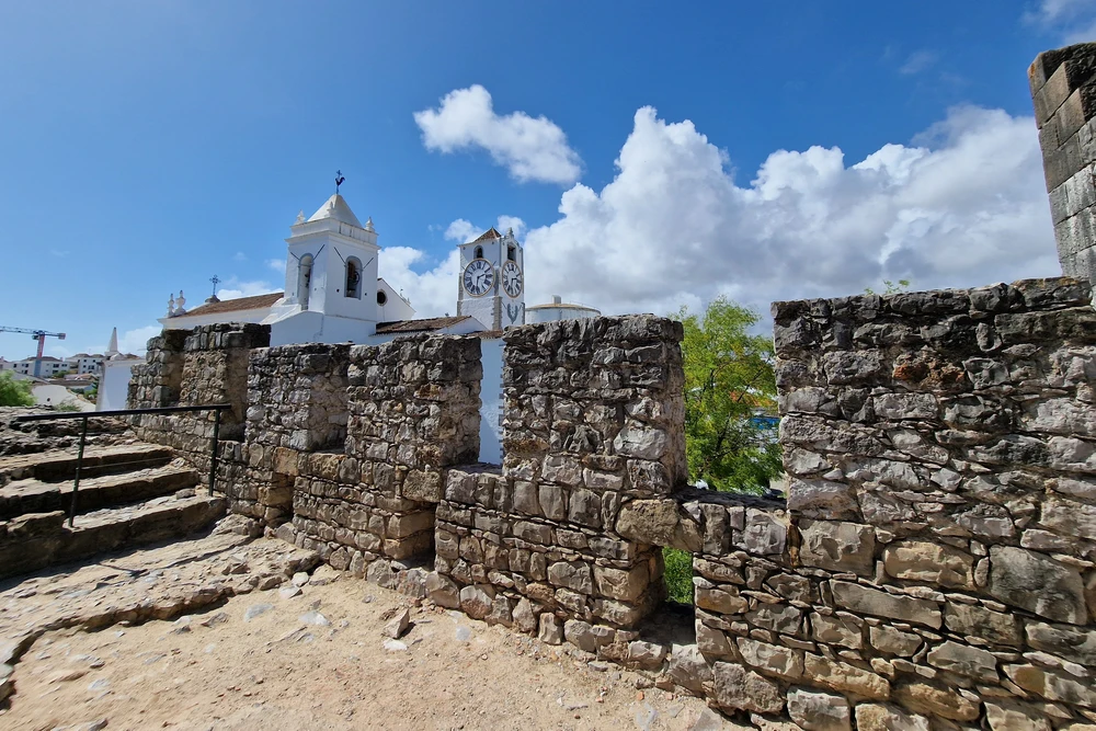 Ancient stone ramparts and walls of the historic Castelo de Tavira, with the white tower of the church visible in the background.