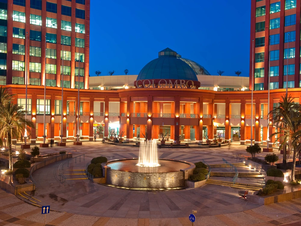 The beautifully illuminated modern facade of the Centro Colombo shopping mall in Lisbon at dusk, with a large fountain in the central plaza.