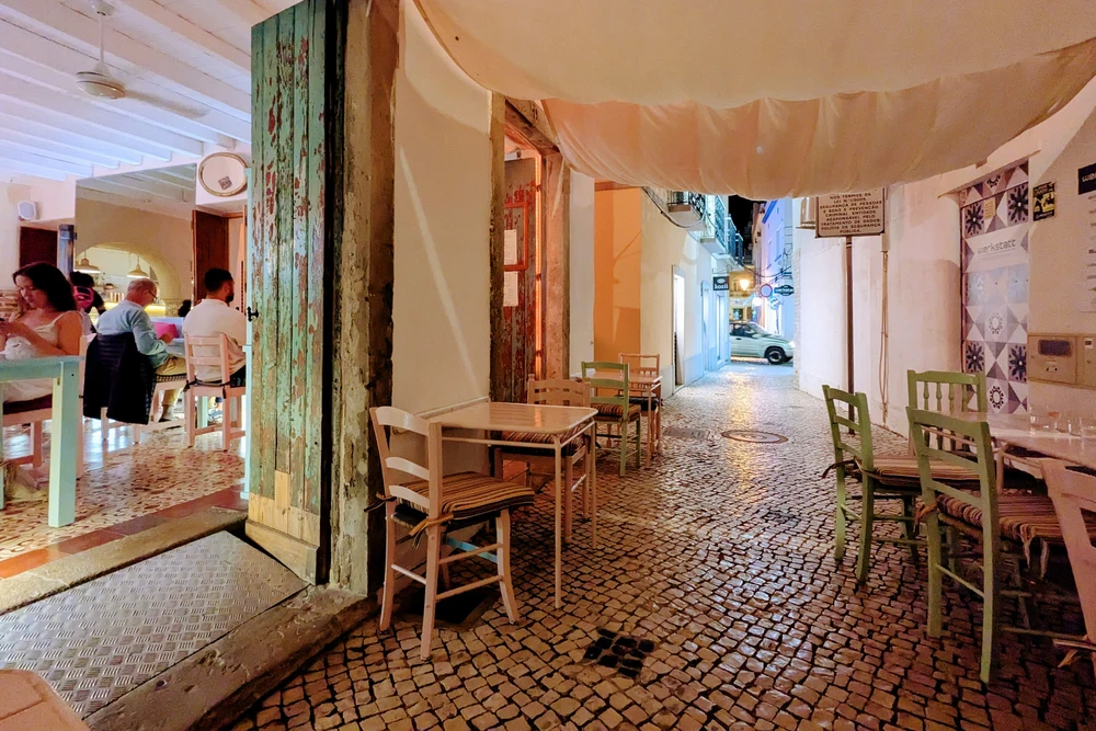 Charming cobblestone alleyway with outdoor seating for a restaurant, featuring rustic wooden chairs and draped fabric in Olhao, Portugal.