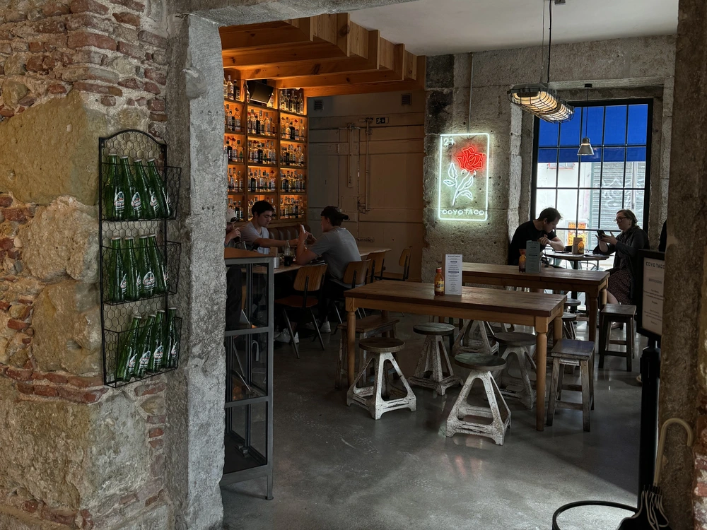 The rustic and trendy interior of the Coyo Taco restaurant in Lisbon, showing patrons dining amidst exposed brick walls and a neon rose sign.