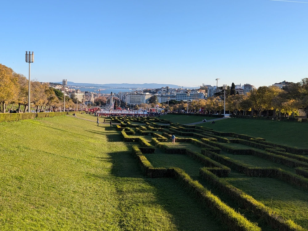 A beautiful panoramic view from the top of Eduardo VII Park in Lisbon, looking down over the geometric hedge patterns and manicured lawns towards the city center.