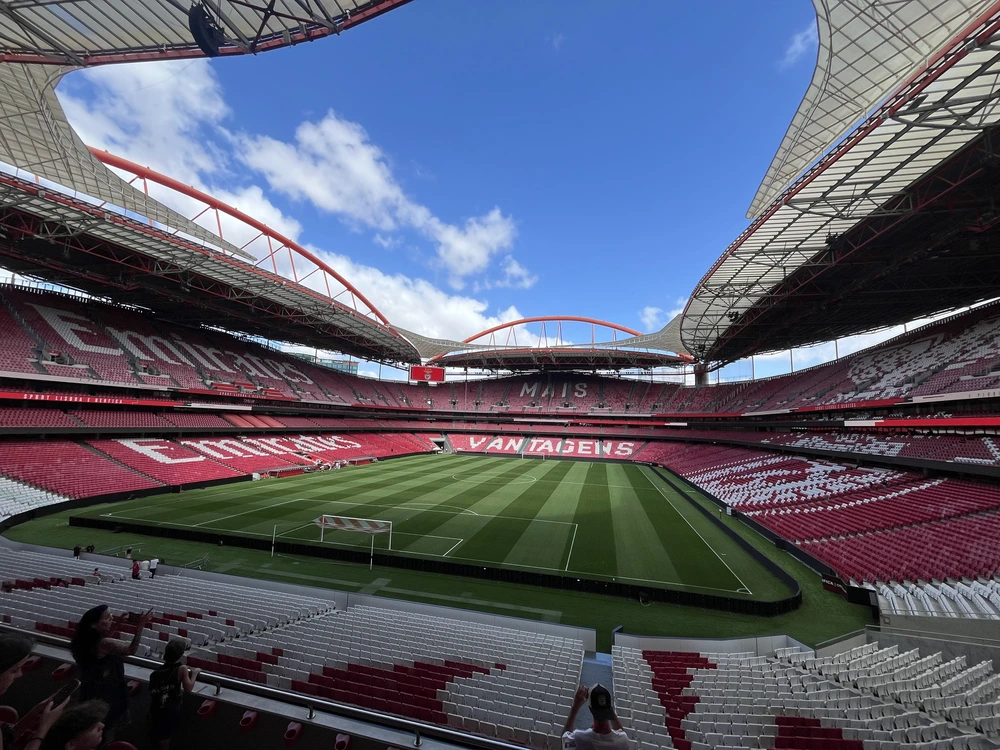 A wide-angle view from the stands of the impressive Estádio da Luz in Lisbon, the home stadium of the Benfica football club, overlooking the vibrant green pitch under a bright blue sky.