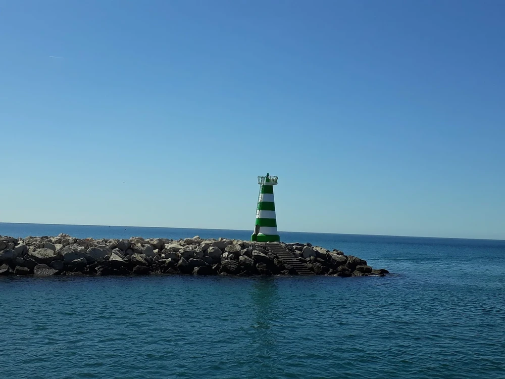 The iconic green and white striped Farol de Vilamoura lighthouse at the end of the marina's rocky jetty on a clear, sunny day.