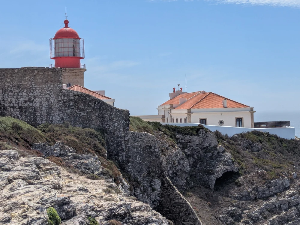 The iconic red-topped lighthouse of Sagres, viewed from behind the ancient stone walls of the fortress on the cliffs.