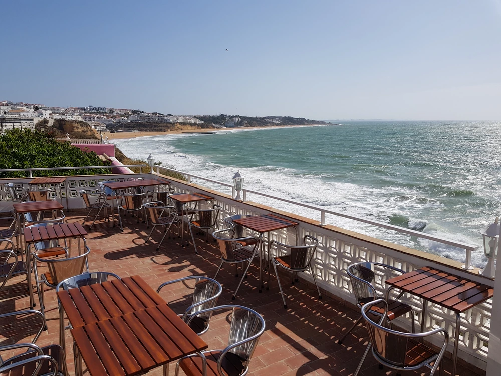 The spectacular coastal view from the sunny terrace of the Iguana Cafe in Albufeira, with tables set for guests to enjoy the crashing waves and ocean scenery.