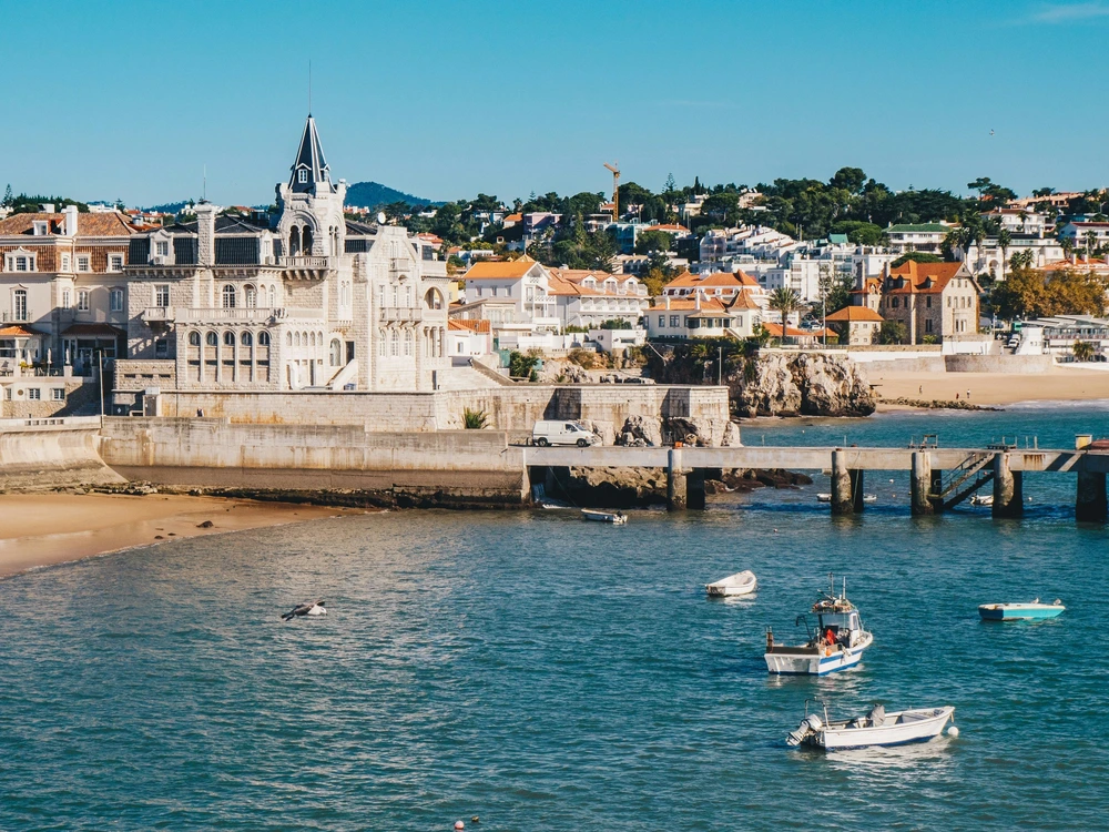 A scenic coastal view of the charming town of Cascais, a popular beach destination for a day trip from Lisbon, with small fishing boats in the turquoise water.