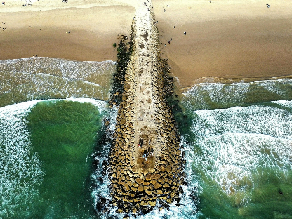 An aerial drone shot of a long stone jetty stretching into the ocean at Costa da Caparica, a popular beach day trip from Lisbon, with waves breaking on either side.