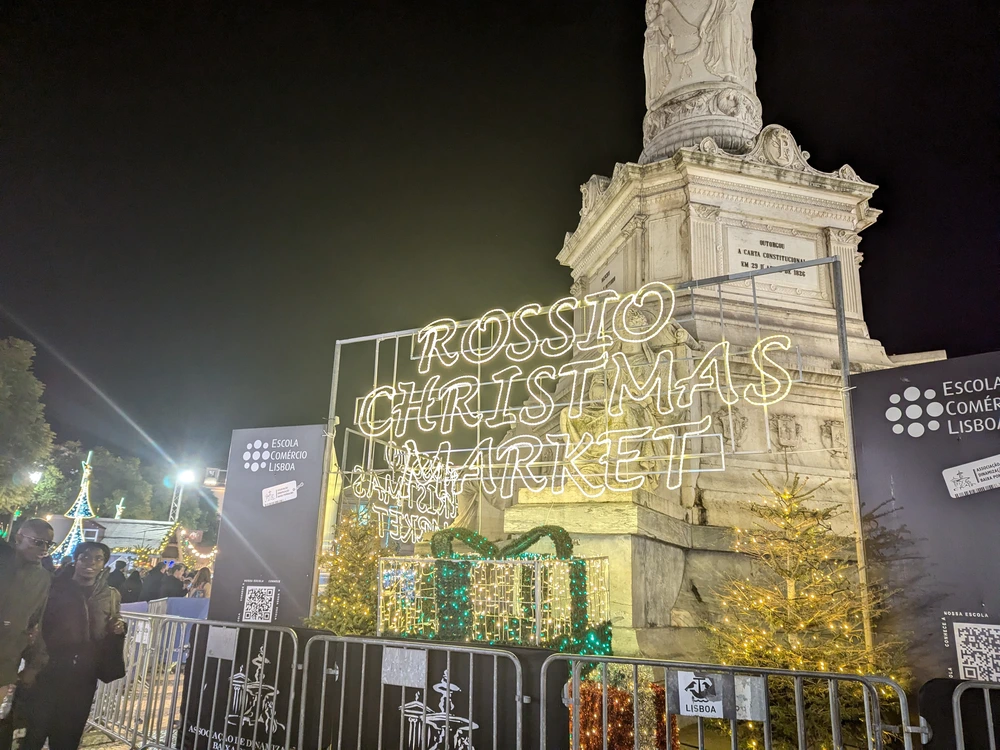 The festive, brightly lit sign for the Rossio Christmas Market in Lisbon at night, with the Column of Pedro IV monument in the background.