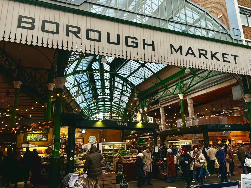 The iconic entrance to Borough Market in London, showing the high glass roof, green ironwork, and bustling crowds of shoppers below.