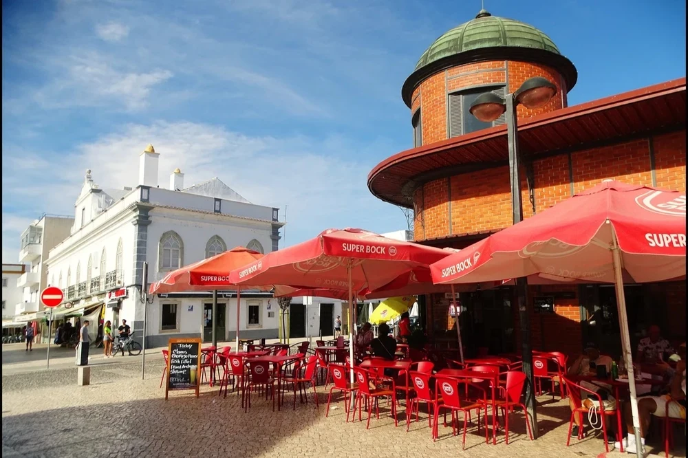 Red brick market building with a domed turret and outdoor seating area with red parasols and chairs in Olhao, Portugal.