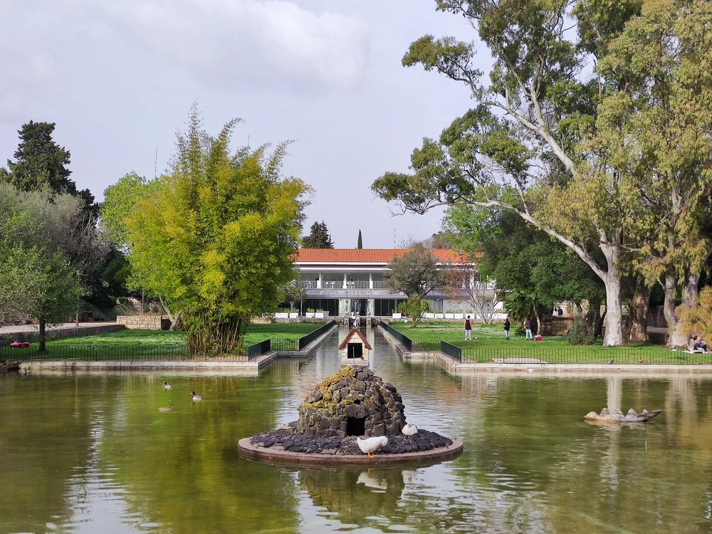 A peaceful scene at Monsanto Forest Park in Lisbon, featuring a tranquil pond with a small duck house and lush green trees in the background.