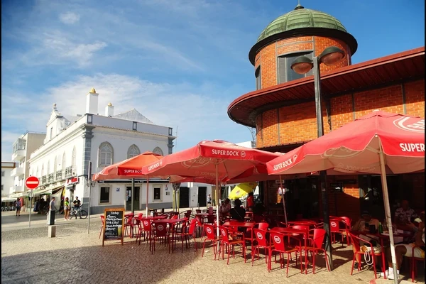Red brick market building with a domed turret and outdoor seating area with red parasols and chairs in Olhao, Portugal.