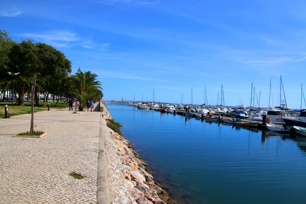 Scenic waterfront promenade lined with palm trees and boats docked along the calm waters of the Olhao, Portugal marina.