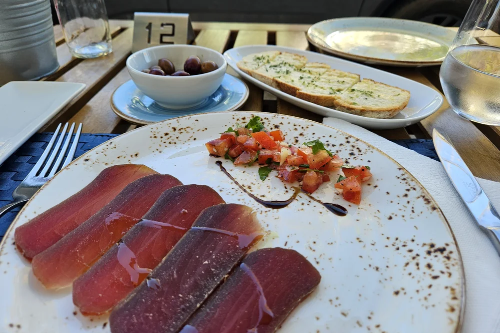 Appetizer platter featuring sliced cured meat, fresh tomato relish, and a side of olives and garlic bread, photographed outdoors at Pausa Restaurant in Tavira.