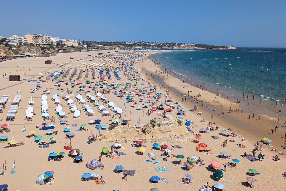 Crowded summer day at Praia da Rocha beach in Portimão, Portugal, with colorful umbrellas, sunbathers, and the Atlantic Ocean shoreline stretching into the distance.