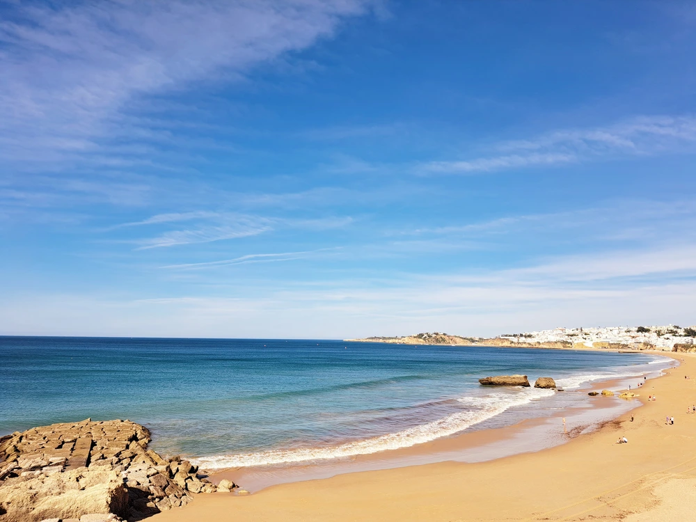 A serene and picturesque view of the expansive Praia de Albufeira, showing golden sand, gentle waves, and the distant white-washed town under a clear blue sky.