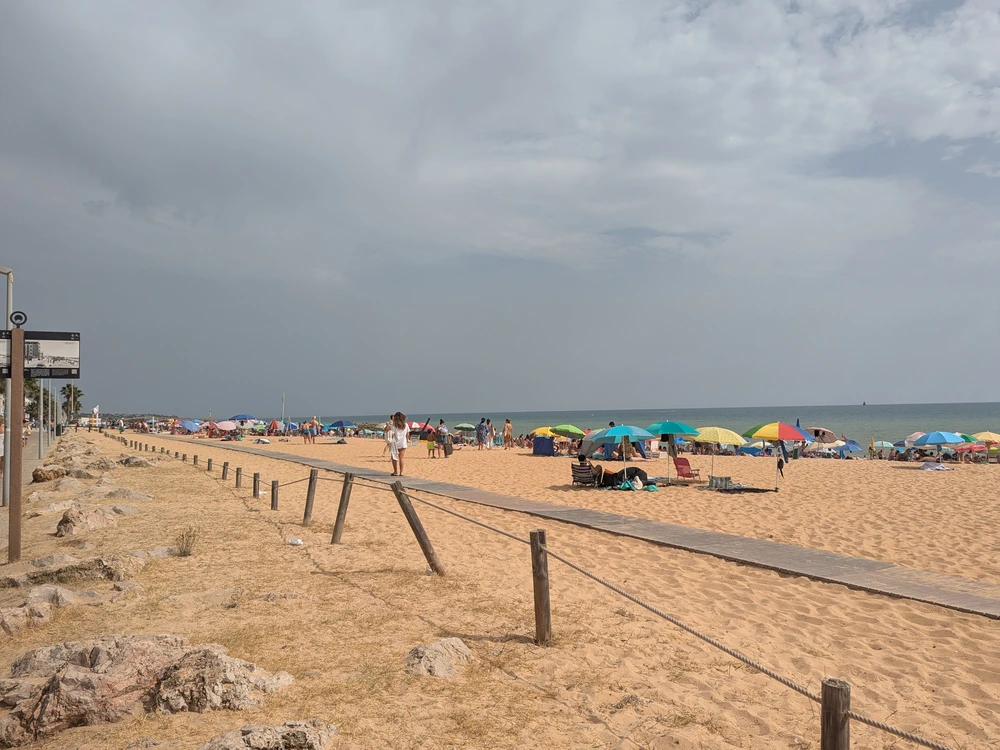 A view across the wide sandy expanse of Praia de Quarteira with a wooden boardwalk and beachgoers in the distance.