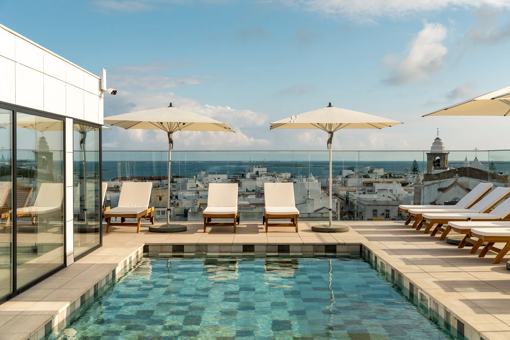 Rooftop terrace pool area with sun loungers overlooking the white buildings and distant church dome of Olhao, Portugal.
