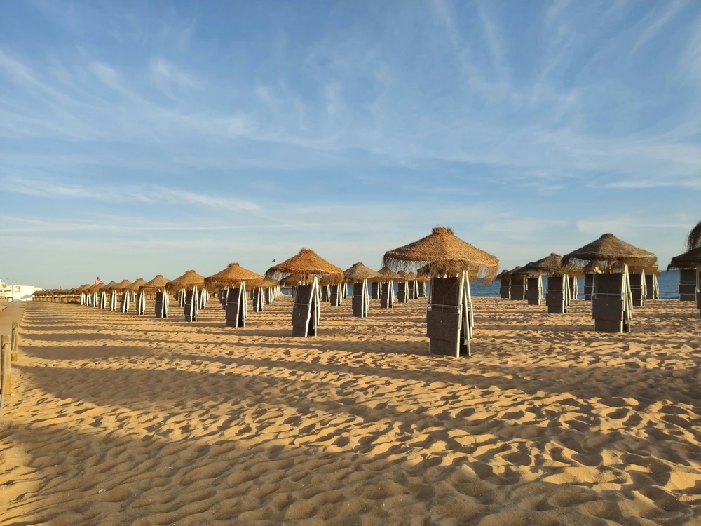 Rows of thatched umbrellas and folded chairs arranged on the golden sand of Quarteira beach in the morning.