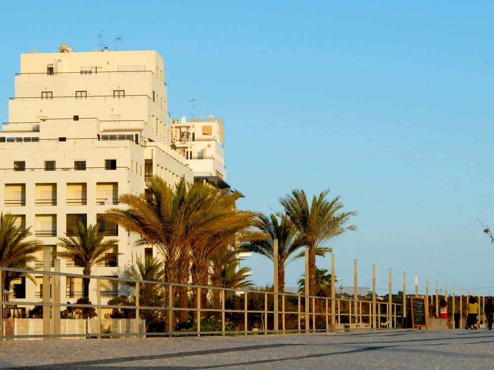 Modern apartment buildings and palm trees along the promenade glow in the sunset light in Quarteira, Portugal.