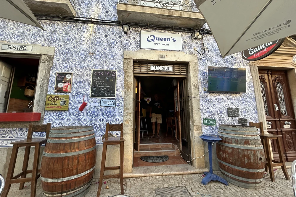 The entrance of Queen's Cafe Sports Bar in Tavira, featuring traditional Portuguese blue and white azulejo tiles and wooden barrels.