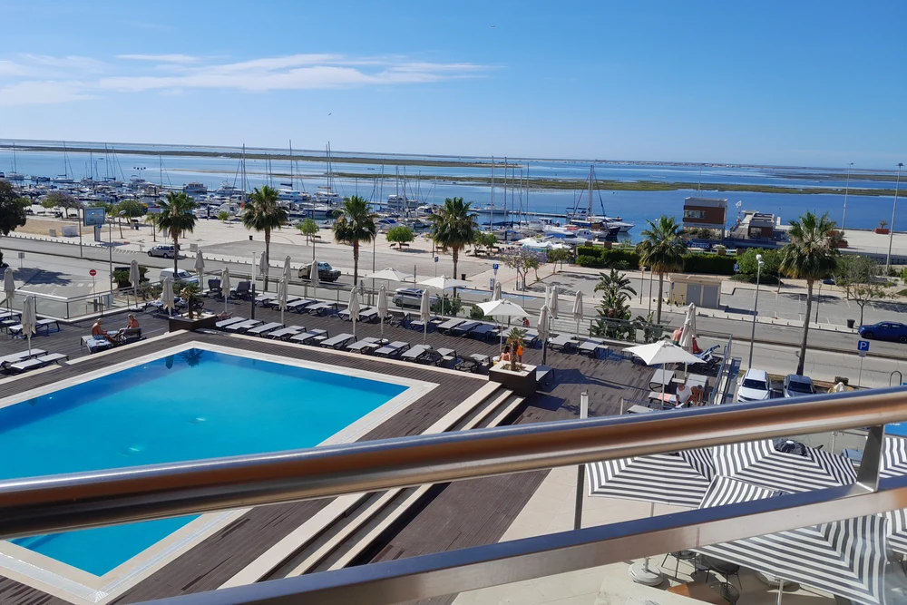 Panoramic view from a balcony showing a swimming pool, palm trees, and the bustling Olhao, Portugal marina.