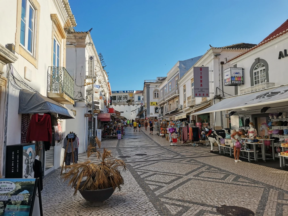 A sunny day on Rua 5 de Outubro, a popular cobblestone shopping street in Old Town Albufeira, with tourists browsing the local shops.