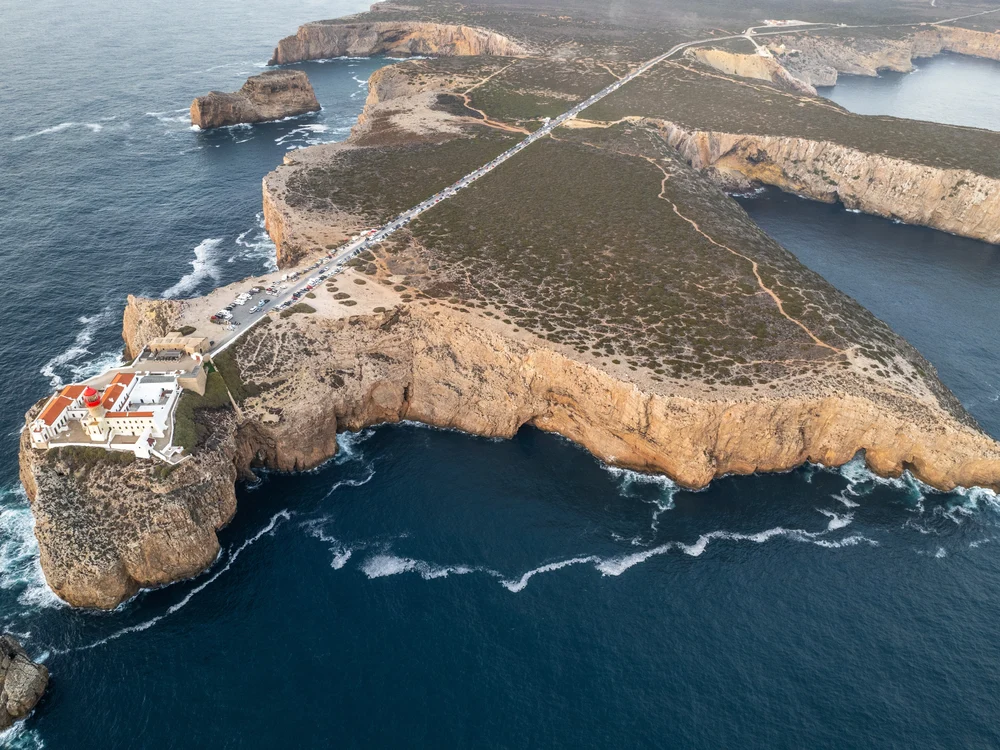A high-angle aerial view of the Cape St. Vincent peninsula in Sagres, with a long road leading to the historic lighthouse complex.