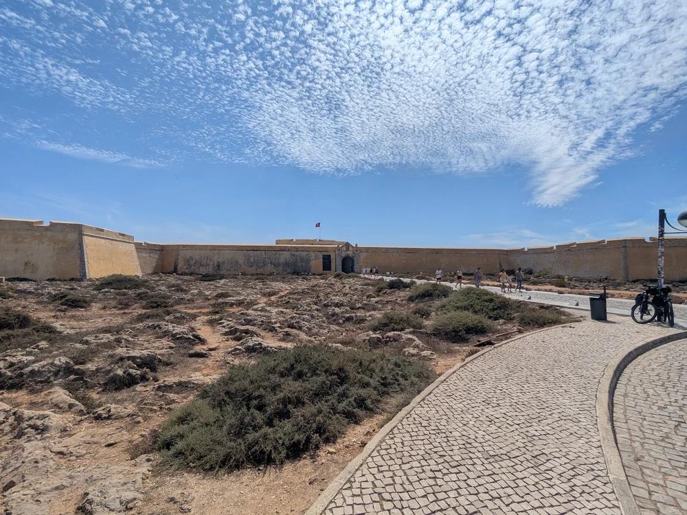 The impressive and historic stone entrance walls of the Fortaleza de Sagres, with a cobblestone path leading visitors inside.