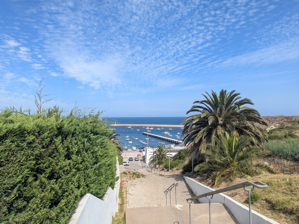 A scenic view from a hilltop in Sagres, looking down a staircase past a palm tree to the tranquil harbor filled with boats.